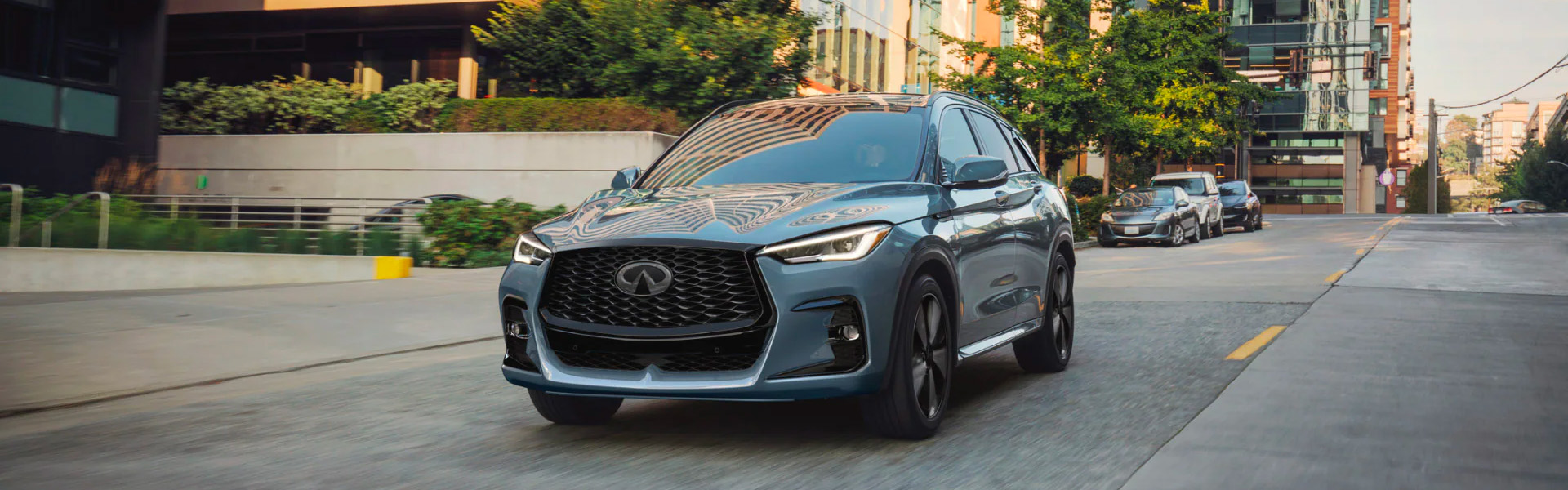 Front view of a light blue 2025 INFINITI QX50 driving down a city street on a sunny day