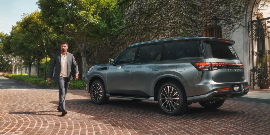 A man in a suit walks confidently on a sunlit cobblestone path beside a sleek gray SUV. Lush trees and a vine-covered wall add elegance to the scene.