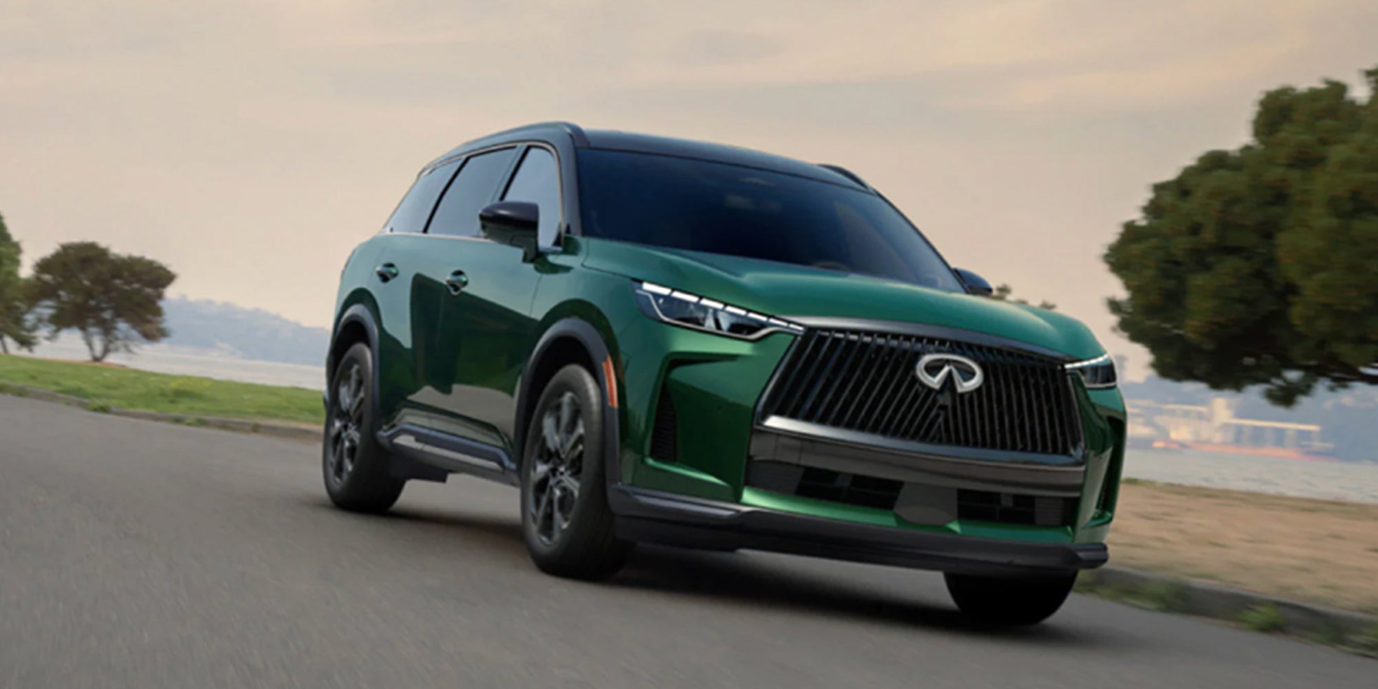 A green 2027 INFINITI QX60 driving along a coastal road with trees and water in the background.